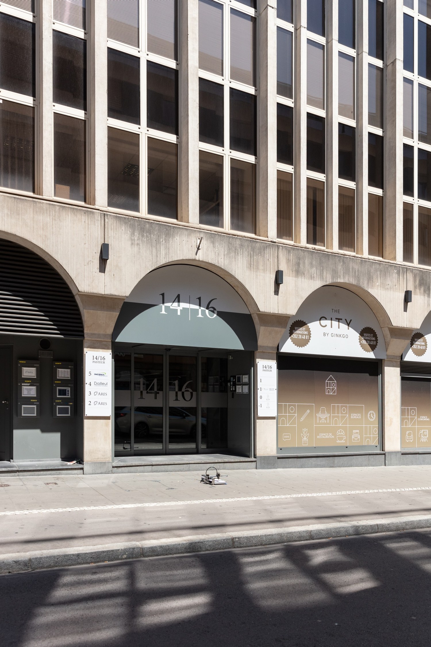14 and 16, Avenue Pasteur, under the arcade. The ground floor is papered with letting graphics for a coworking operator, a directory panel beside the door lists the tenants of the floors above, and the plotter draws on the pavement outside.