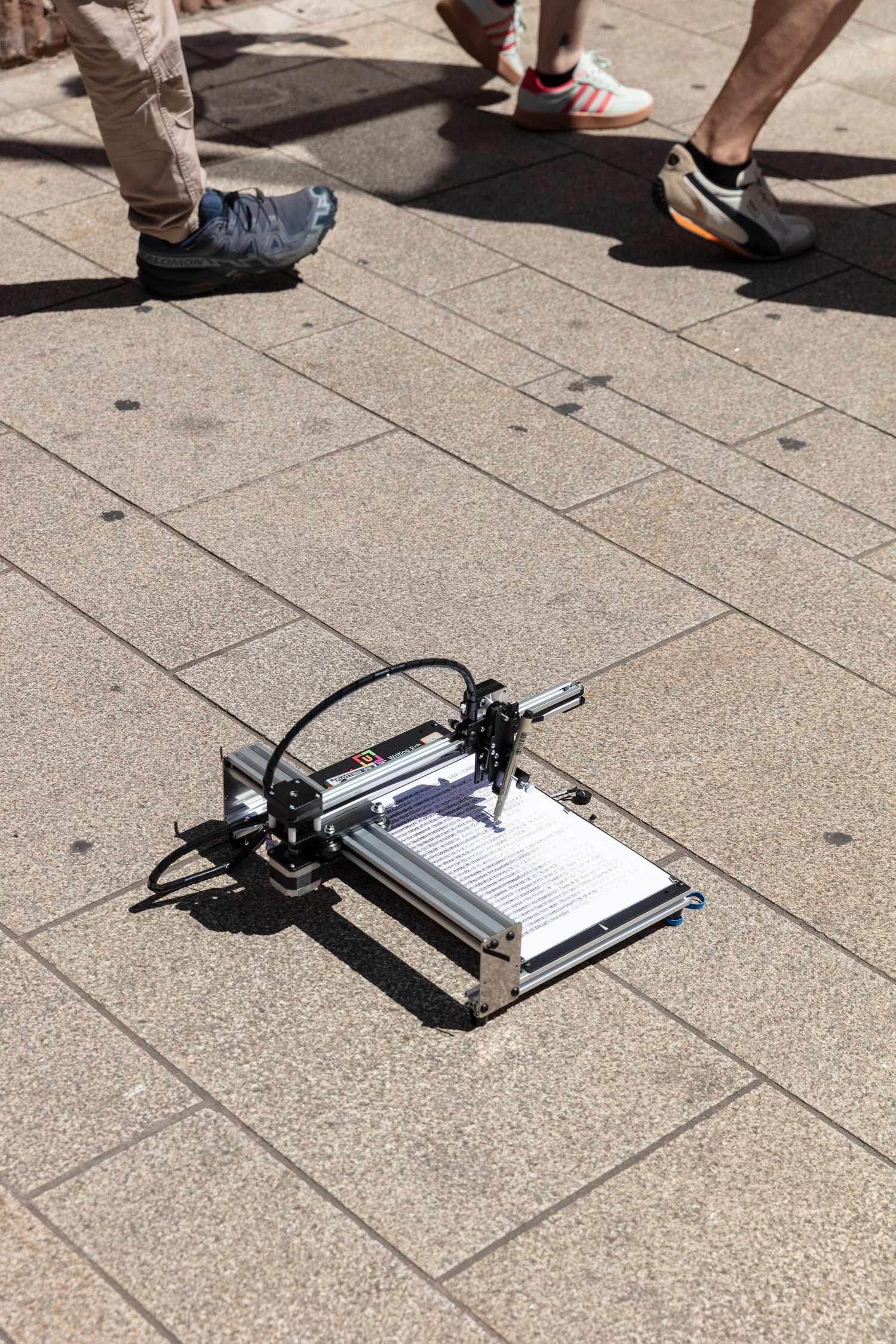 The plotter at work on the pavement, drawing the sheet for the address it stands in front of, the page half written as pedestrians pass without stopping.
